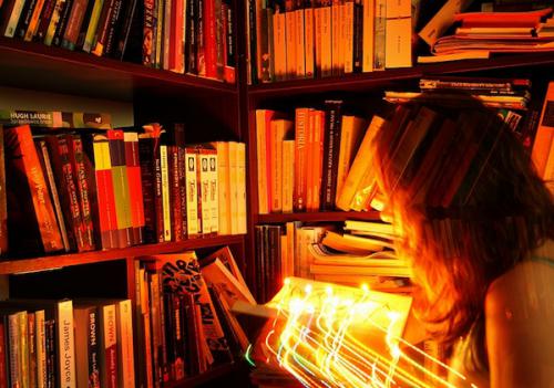 Hidden Fire Standing among bookshelves, a woman holds an open book with bright lights shooting out of it